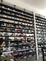 A shoe store wall filled with multiple rows of sneakers, displaying a variety of brands, styles, and colors. The sneakers are arranged neatly on black shelves against a white wall. There are hints of other merchandise and clothing visible in the background.