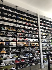 A shoe store wall filled with multiple rows of sneakers, displaying a variety of brands, styles, and colors. The sneakers are arranged neatly on black shelves against a white wall. There are hints of other merchandise and clothing visible in the background.