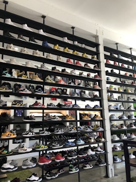 A shoe store wall filled with multiple rows of sneakers, displaying a variety of brands, styles, and colors. The sneakers are arranged neatly on black shelves against a white wall. There are hints of other merchandise and clothing visible in the background.