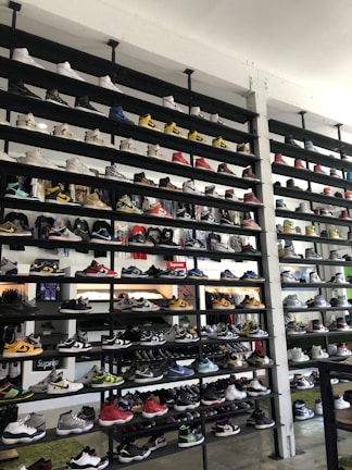 A shoe store wall filled with multiple rows of sneakers, displaying a variety of brands, styles, and colors. The sneakers are arranged neatly on black shelves against a white wall. There are hints of other merchandise and clothing visible in the background.