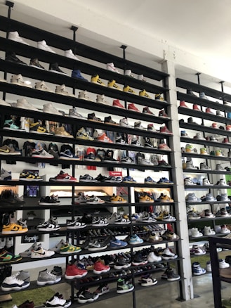 A shoe store wall filled with multiple rows of sneakers, displaying a variety of brands, styles, and colors. The sneakers are arranged neatly on black shelves against a white wall. There are hints of other merchandise and clothing visible in the background.