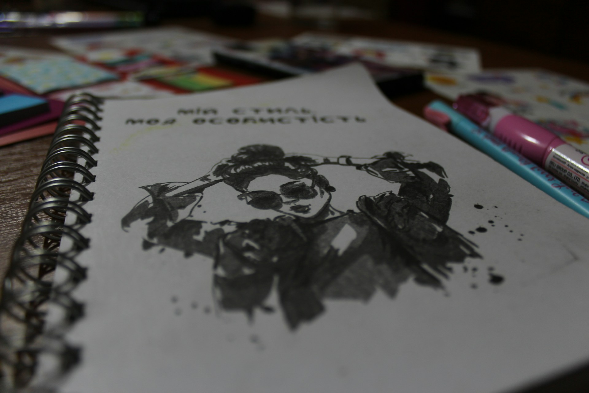 A spiral-bound notebook is open to a sketch featuring a stylized ink drawing of a person wearing sunglasses and a jacket. The drawing includes splatter effects for a dynamic look. Around the notebook, various colored pens and art supplies are scattered, slightly out of focus.