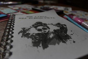 A spiral-bound notebook is open to a sketch featuring a stylized ink drawing of a person wearing sunglasses and a jacket. The drawing includes splatter effects for a dynamic look. Around the notebook, various colored pens and art supplies are scattered, slightly out of focus.