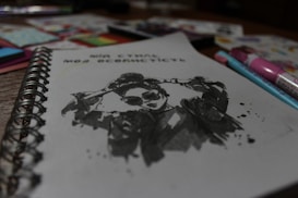 A spiral-bound notebook is open to a sketch featuring a stylized ink drawing of a person wearing sunglasses and a jacket. The drawing includes splatter effects for a dynamic look. Around the notebook, various colored pens and art supplies are scattered, slightly out of focus.