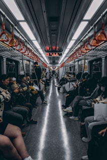 A crowded subway car with passengers seated on both sides. Many people are engrossed in their phones, and the interior features overhead handrails with orange grips. The scene is lit with bright fluorescent lights, and a digital sign displays information above.