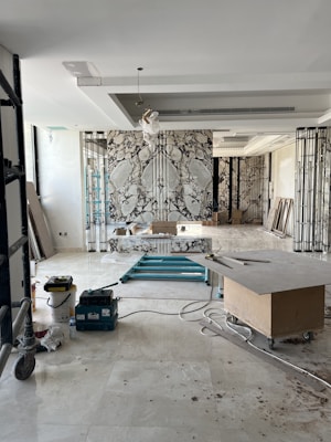 An interior space appears to be under renovation, featuring a large, decorative marble wall with an intricate pattern. Scaffolding, tools, and materials are scattered throughout the room, including a cart, power tools, and a table. The room is lit by natural light coming through large windows, adding to the sense of openness.