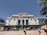 A historic train station with a prominent 'Union Station' sign in orange letters on its facade. The architecture features classical elements with arched windows and decorative stonework. In the foreground, an American flag is prominently displayed on a flagpole. People are walking and cycling in the area, and there are outdoor tables with umbrellas near the station.