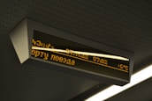 A digital display board is mounted on the ceiling. It features amber-colored text in a foreign language that includes numbers, possibly representing time or departure information. The board is encased in a rectangular frame and is part of a public transportation environment.