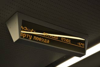 A digital display board is mounted on the ceiling. It features amber-colored text in a foreign language that includes numbers, possibly representing time or departure information. The board is encased in a rectangular frame and is part of a public transportation environment.