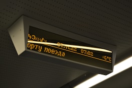 A digital display board is mounted on the ceiling. It features amber-colored text in a foreign language that includes numbers, possibly representing time or departure information. The board is encased in a rectangular frame and is part of a public transportation environment.