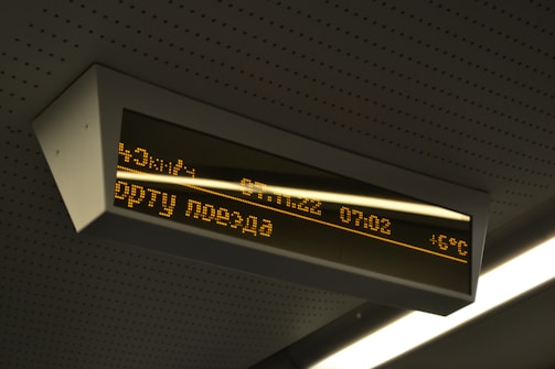 A digital display board is mounted on the ceiling. It features amber-colored text in a foreign language that includes numbers, possibly representing time or departure information. The board is encased in a rectangular frame and is part of a public transportation environment.