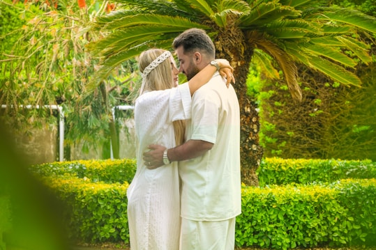 A couple embraces warmly in a lush garden, surrounded by vibrant green foliage and a large palm-like tree. Both individuals are dressed in white, evoking a serene and harmonious vibe amidst nature.