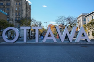 Large block letters spelling 'Ottawa' are prominently displayed in front of historic stone buildings and bare trees with autumn foliage in the background. The scene includes a clear blue sky with a few clouds, and a construction crane is visible in the distance.