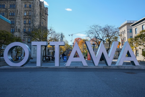 Large block letters spelling 'Ottawa' are prominently displayed in front of historic stone buildings and bare trees with autumn foliage in the background. The scene includes a clear blue sky with a few clouds, and a construction crane is visible in the distance.