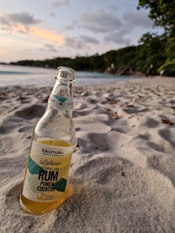 A bottle of Takamaka sparkling rum punch cocktail is partially buried in sandy beach, with a calm ocean and distant green trees in the background. The sky is partly cloudy with hints of pink and orange from the setting sun.