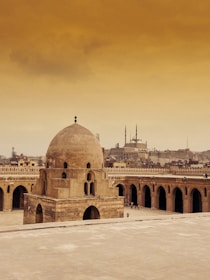 A historical mosque with large domes and intricate architecture is set against a city skyline. The mosque features tall minarets and a vast courtyard surrounded by arches. The sky above is a warm, golden hue, adding to the ancient and tranquil atmosphere.