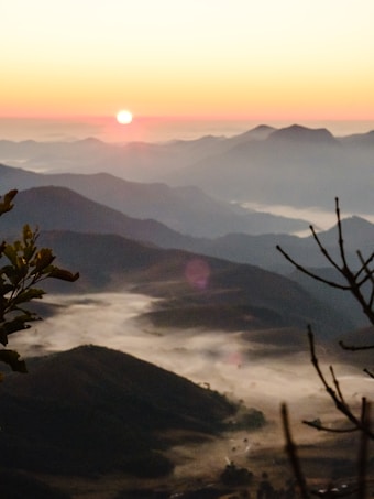 The image captures a tranquil mountain landscape at sunrise. The sun is rising over distant mountain ranges, casting a warm, orange and pink hue across the sky. Mist and fog linger in the valleys between the dark silhouettes of the hills, creating a serene and peaceful ambiance. In the foreground, branches of a tree partially frame the scene.