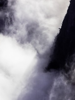 Thick, swirling clouds envelop a dark rocky cliff, creating a dramatic contrast between the misty white vapors and the shadowy landscape. The scene is mysterious and atmospheric, with clouds possibly indicating weather changes or elevation.