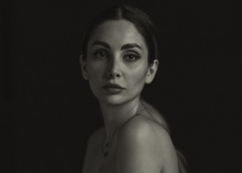 A monochromatic portrait of a woman with a neutral expression, gazing directly at the camera. The image is dark and appears to be taken in a low-light setting, with subtle highlights on the woman's face and shoulders. Her hair is pulled back, and there is a minimalistic and artistic feel to the composition.