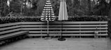 A wooden deck with two closed patio umbrellas standing upright. One umbrella has black and white stripes, while the other is a solid color. The deck is surrounded by a wooden railing with a hedge behind it. In the background, there are numerous trees, creating a forest-like setting. A bench is built into one side of the deck, and there are small scattered leaves on the wooden floor.