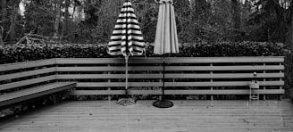 A wooden deck with two closed patio umbrellas standing upright. One umbrella has black and white stripes, while the other is a solid color. The deck is surrounded by a wooden railing with a hedge behind it. In the background, there are numerous trees, creating a forest-like setting. A bench is built into one side of the deck, and there are small scattered leaves on the wooden floor.