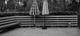 A wooden deck with two closed patio umbrellas standing upright. One umbrella has black and white stripes, while the other is a solid color. The deck is surrounded by a wooden railing with a hedge behind it. In the background, there are numerous trees, creating a forest-like setting. A bench is built into one side of the deck, and there are small scattered leaves on the wooden floor.