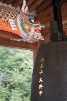 A decorative, colorful dragon head, crafted with intricate detail, appears poised beside a large, dark bronze bell adorned with golden inscriptions. The setting includes a wooden structure and lush green foliage in the background.