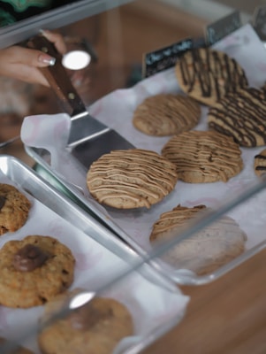 A variety of freshly baked cookies displayed in trays with a hand holding a wooden-handled spatula. The cookies are drizzled with chocolate and have different textures and toppings.