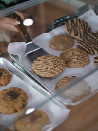 A variety of freshly baked cookies displayed in trays with a hand holding a wooden-handled spatula. The cookies are drizzled with chocolate and have different textures and toppings.