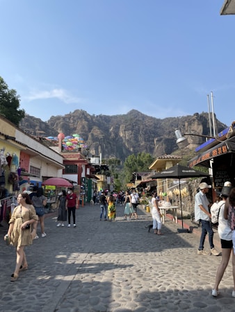A lively street scene with people strolling along a cobblestone path lined with small shops on either side. The area is vibrant with various stalls displaying colorful merchandise. Towering mountains form a scenic backdrop under a clear blue sky. Umbrellas hang above one of the shops, adding a splash of color.