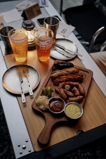 A wooden board on a table holds a variety of sausages with small dishes of ketchup and mustard, along with sauerkraut and pickles. On the same table, two glasses filled with beer are placed next to a small lantern. Metal cutlery is seen on a small round plate beside the wooden board.