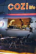 An outdoor food stall with a large neon sign reading 'COZI life' illuminates the scene. Inside the stall, a worker is preparing food, surrounded by kitchen equipment. In front of the stall, several folding tables and chairs are set up for customers. The overall ambiance is cozy and inviting, with a warm glow from the lights.