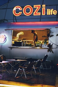An outdoor food stall with a large neon sign reading 'COZI life' illuminates the scene. Inside the stall, a worker is preparing food, surrounded by kitchen equipment. In front of the stall, several folding tables and chairs are set up for customers. The overall ambiance is cozy and inviting, with a warm glow from the lights.