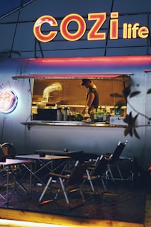 An outdoor food stall with a large neon sign reading 'COZI life' illuminates the scene. Inside the stall, a worker is preparing food, surrounded by kitchen equipment. In front of the stall, several folding tables and chairs are set up for customers. The overall ambiance is cozy and inviting, with a warm glow from the lights.