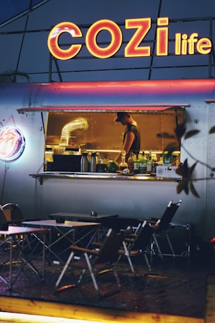 An outdoor food stall with a large neon sign reading 'COZI life' illuminates the scene. Inside the stall, a worker is preparing food, surrounded by kitchen equipment. In front of the stall, several folding tables and chairs are set up for customers. The overall ambiance is cozy and inviting, with a warm glow from the lights.