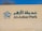 The image features a beige wall with green lettering spelling 'Al-Azhar Park' alongside Arabic script. A stylized floral logo in green and white is positioned to the left of the text. Part of a tree is visible above the wall against a clear blue sky.