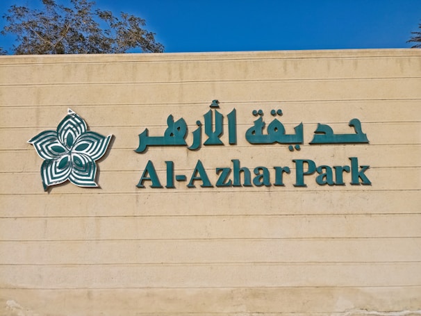The image features a beige wall with green lettering spelling 'Al-Azhar Park' alongside Arabic script. A stylized floral logo in green and white is positioned to the left of the text. Part of a tree is visible above the wall against a clear blue sky.