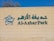 The image features a beige wall with green lettering spelling 'Al-Azhar Park' alongside Arabic script. A stylized floral logo in green and white is positioned to the left of the text. Part of a tree is visible above the wall against a clear blue sky.