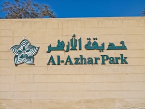 The image features a beige wall with green lettering spelling 'Al-Azhar Park' alongside Arabic script. A stylized floral logo in green and white is positioned to the left of the text. Part of a tree is visible above the wall against a clear blue sky.