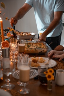 A cozy dining setting with a table filled with elegant dinnerware and a variety of foods. A person is serving food from a casserole dish while another prepares to eat. Decorative elements include candles and small floral arrangements, creating a warm and inviting atmosphere.