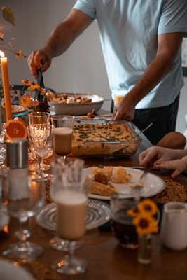 A cozy dining setting with a table filled with elegant dinnerware and a variety of foods. A person is serving food from a casserole dish while another prepares to eat. Decorative elements include candles and small floral arrangements, creating a warm and inviting atmosphere.