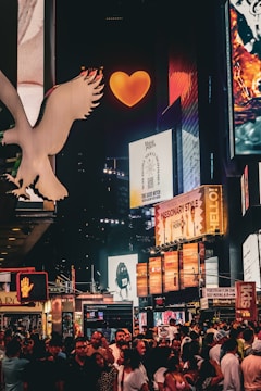 A vibrant nighttime city scene filled with illuminated billboards and advertisements. The atmosphere buzzes with a crowd of people on the street, while bright signage and logos surround them. A large, glowing orange heart captures attention at the top center, amidst numerous colorful digital displays.