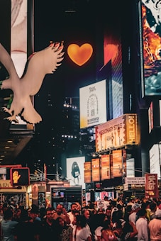 A vibrant nighttime city scene filled with illuminated billboards and advertisements. The atmosphere buzzes with a crowd of people on the street, while bright signage and logos surround them. A large, glowing orange heart captures attention at the top center, amidst numerous colorful digital displays.