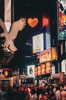 A vibrant nighttime city scene filled with illuminated billboards and advertisements. The atmosphere buzzes with a crowd of people on the street, while bright signage and logos surround them. A large, glowing orange heart captures attention at the top center, amidst numerous colorful digital displays.