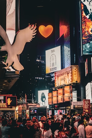 A vibrant nighttime city scene filled with illuminated billboards and advertisements. The atmosphere buzzes with a crowd of people on the street, while bright signage and logos surround them. A large, glowing orange heart captures attention at the top center, amidst numerous colorful digital displays.