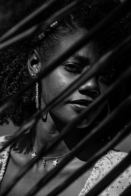 A black and white portrait of a person with braided hair, partially obscured by shadows cast by what appears to be plant leaves. The person is wearing hoop earrings and a necklace with small floral pendants. The lighting creates a dramatic play of light and dark across their face.