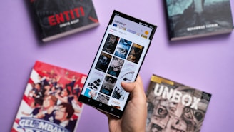 A hand holding a smartphone displaying a digital library or reading app interface, surrounded by several books with colorful covers. The background is a solid light purple surface.