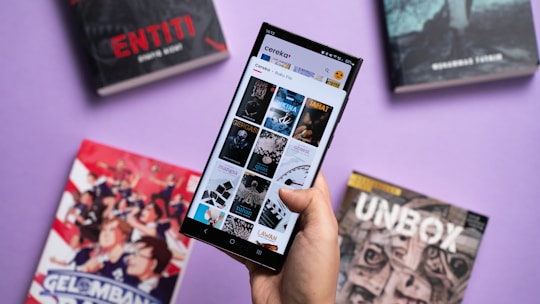 A hand holding a smartphone displaying a digital library or reading app interface, surrounded by several books with colorful covers. The background is a solid light purple surface.