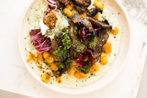 A plate contains a delicious arrangement of food, featuring roasted mushrooms, crispy gnocchi, and vibrant radicchio leaves. The dish is topped with a green herb garnish, likely parsley or cilantro, and rests on a creamy green sauce. The colors are vivid and fresh.