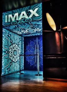 An entrance with ornate blue and teal designs covers the walls and ceiling, featuring intricate floral patterns. The brand 'IMAX' is prominently displayed at the top in bold white letters. A metallic barrier is present at the entryway, and ambient lighting creates a modern cinematic ambiance.
